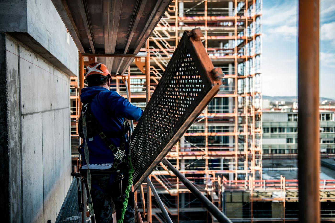 Gerüstbauer mit Auffanggurt trägt einen Gerüstbelag, im Hintergrund ein eingerüsteter Neubau